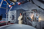 A scientist poses beside an enormous vertical shaft underneath instrumentation.